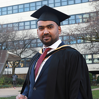 A student wearing a black graduation gown and black graduation cap smiles at the camera outside of a university building with overcast skies.