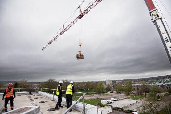 Two people on a roof watch as a crane above them delivers a large wooden box onto the roof